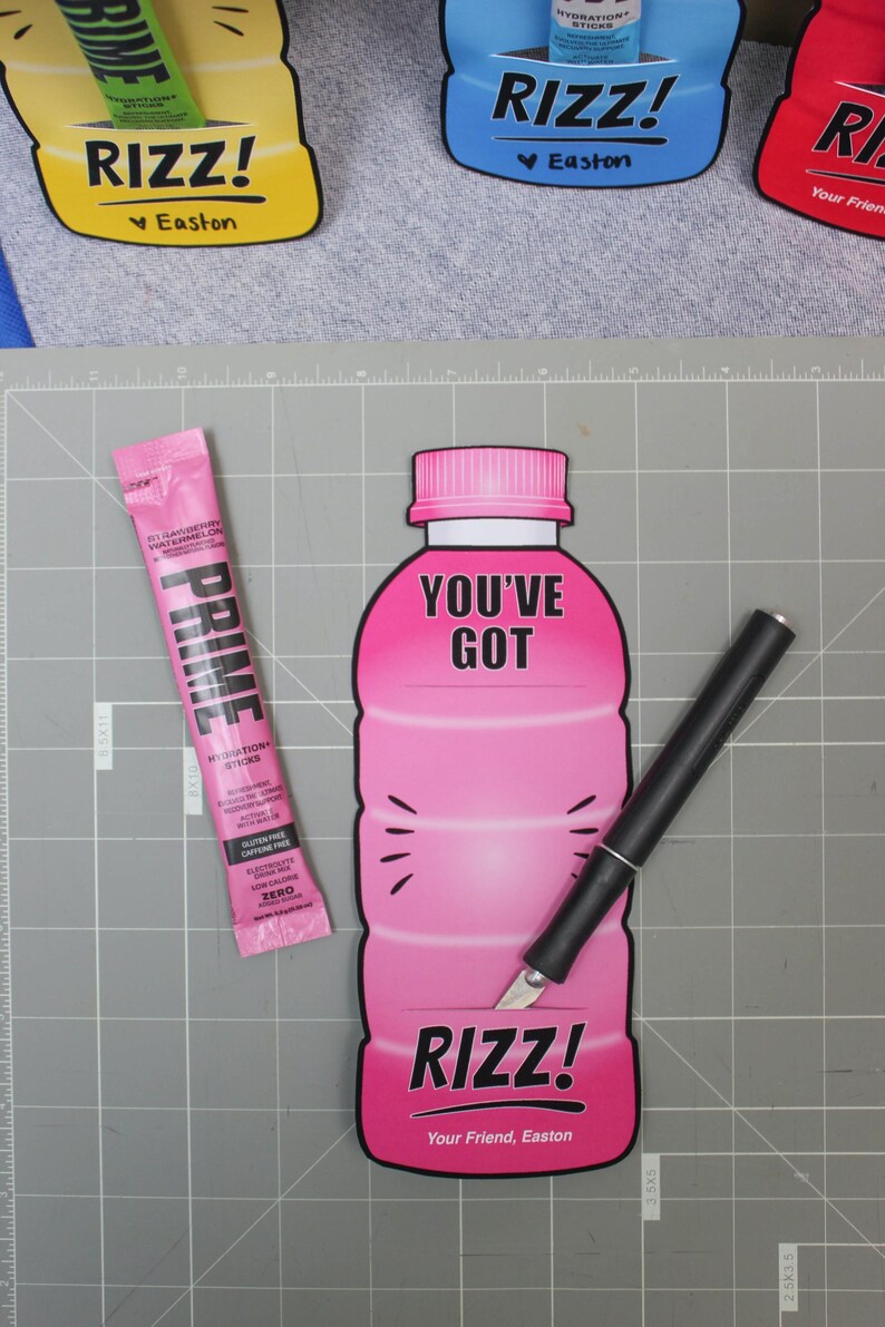 May include: A pink and white cardboard cutout of a sports drink bottle with the text "You've Got Rizz! Your Friend, Easton" on it. A black utility knife is cutting a slit in the bottle to insert a pink and white Prime Hydration stick.