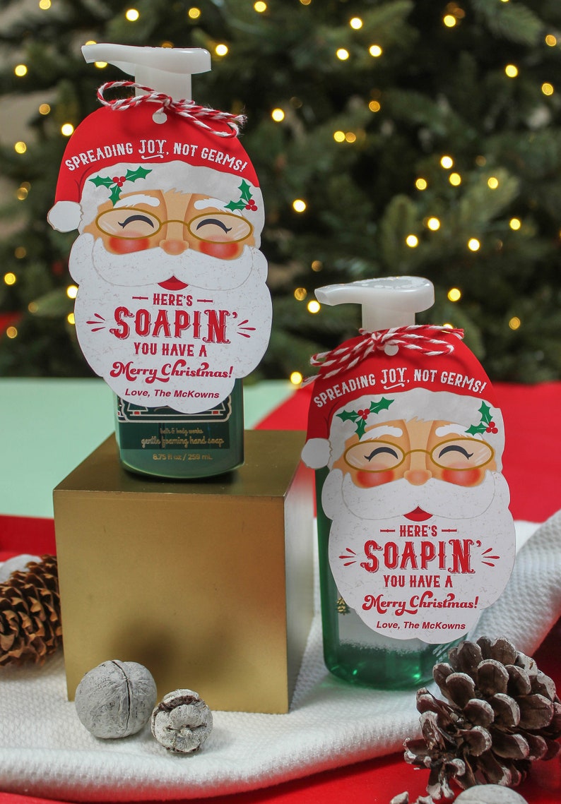 Op de afbeelding: Twee groene flessen met schuimende handzeep met kerstman-cadeautjeslabels. De labels zeggen "Here's Soapin' You Have a Merry Christmas! Love, The McKowns" en "Spreading Joy, Not Germs!"