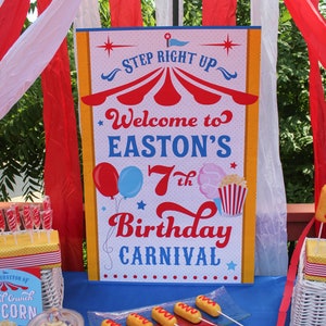 Peut inclure: Mise en place pour une f&ecirc;te d'anniversaire sur le th&egrave;me du carnaval. Une pancarte indique "Bienvenue au 7e anniversaire de Easton" avec des ballons, du pop-corn et des illustrations de barbe &agrave; papa. Des hot-dogs, des cupcakes et du pop-corn sont expos&eacute;s.