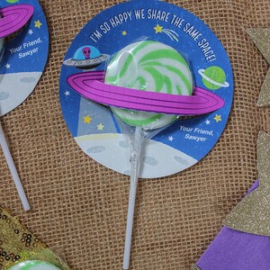 Op de afbeelding: Een groene en witte swirl-lolli met een paarse ring eromheen, bevestigd aan een witte stok. De lolly bevindt zich op een ronde kaart met een blauwe achtergrond en een cartoon-alien-ruimteschip. De kaart zegt "Ik ben zo blij dat we dezelfde ruimte delen!" en "Je vriend, Sawyer".
