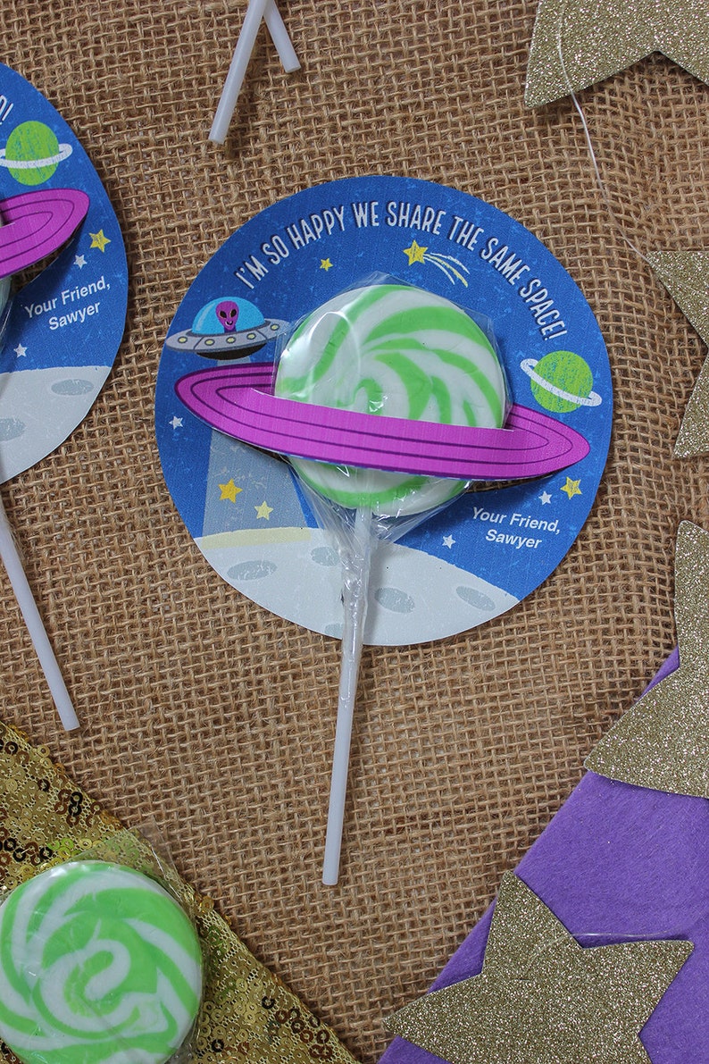Op de afbeelding: Een groene en witte swirl-lolli met een paarse ring eromheen, bevestigd aan een witte stok. De lolly bevindt zich op een ronde kaart met een blauwe achtergrond en een cartoon-alien-ruimteschip. De kaart zegt "Ik ben zo blij dat we dezelfde ruimte delen!" en "Je vriend, Sawyer".