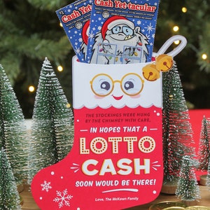 Pode incluir: Uma meia vermelha com uma bainha branca e um rosto branco com &oacute;culos. A meia est&aacute; cheia de dois bilhetes de loteria que dizem "Cash Yet-tacular". A meia est&aacute; sentada sobre uma superf&iacute;cie de madeira com duas pequenas &aacute;rvores de pinho e moedas de ouro.