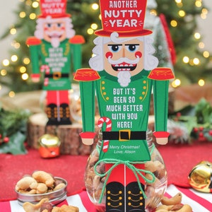 May include: A nutcracker figurine holding a jar of mixed nuts. The nutcracker is green and red with a white face and black mustache. The jar is clear glass and has a red ribbon tied around it. The text on the nutcracker reads "Another Nutty Year" and "But it's been so much better with you here!"  Merry Christmas! Love, Alison