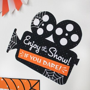 May include: Black and white Halloween party sign shaped like a vintage movie camera with the text "Enjoy the Show! If you dare!" in white lettering on a black background.