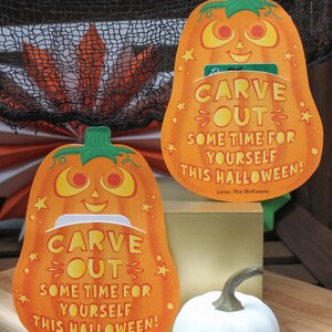 May include: Two orange pumpkin-shaped cards with a cut-out slot in the middle. The cards have the text "CARVE OUT SOME TIME FOR YOURSELF THIS HALLOWEEN! Love, The McKowns" on them. There are two white pumpkins in the background.
