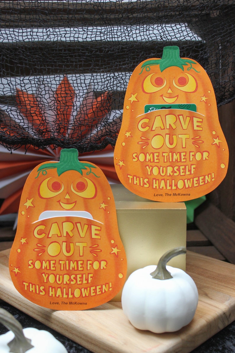 May include: Two orange pumpkin-shaped cards with a cut-out slot in the middle. The cards have the text "CARVE OUT SOME TIME FOR YOURSELF THIS HALLOWEEN! Love, The McKowns" on them. There are two white pumpkins in the background.