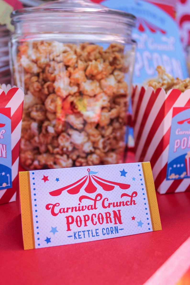 May include: A white sign with red, yellow, and blue accents reads "Carnival Crunch Popcorn - Kettle Corn" with a red and white striped tent graphic. The sign is on a red surface with a glass jar of popcorn in the background.