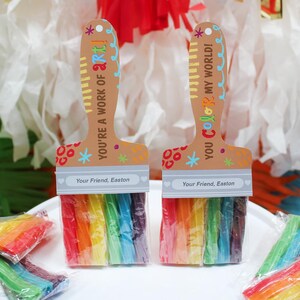 May include: Two paint brush-shaped party favors with rainbow-colored candy bristles. The handles are tan with colorful text that reads "YOU'RE A WORK OF ART!" and "YOU COLOR MY WORLD!" The text "Your Friend, Easton" is printed on the bottom. Several wrapped candy bundles are scattered around.