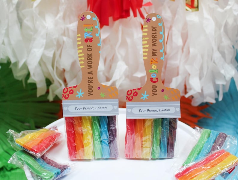 May include: Two paint brush-shaped party favors with rainbow-colored candy bristles. The handles are tan with colorful text that reads "YOU'RE A WORK OF ART!" and "YOU COLOR MY WORLD!" The text "Your Friend, Easton" is printed on the bottom. Several wrapped candy bundles are scattered around.