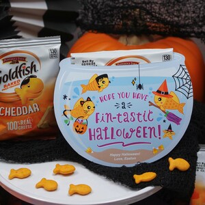 May include: A blue and white Halloween-themed goldfish bowl with a spider web design and the text "Hope you have a fin-tastic Halloween!"  The bowl is surrounded by a black mesh fabric and two boxes of Goldfish crackers.