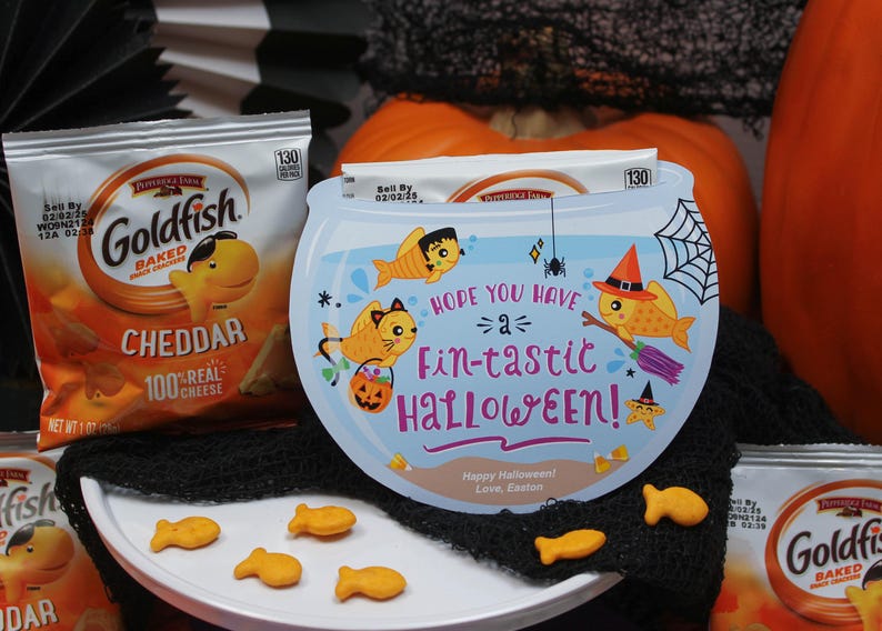 May include: A blue and white Halloween-themed goldfish bowl with a spider web design and the text "Hope you have a fin-tastic Halloween!"  The bowl is surrounded by a black mesh fabric and two boxes of Goldfish crackers.