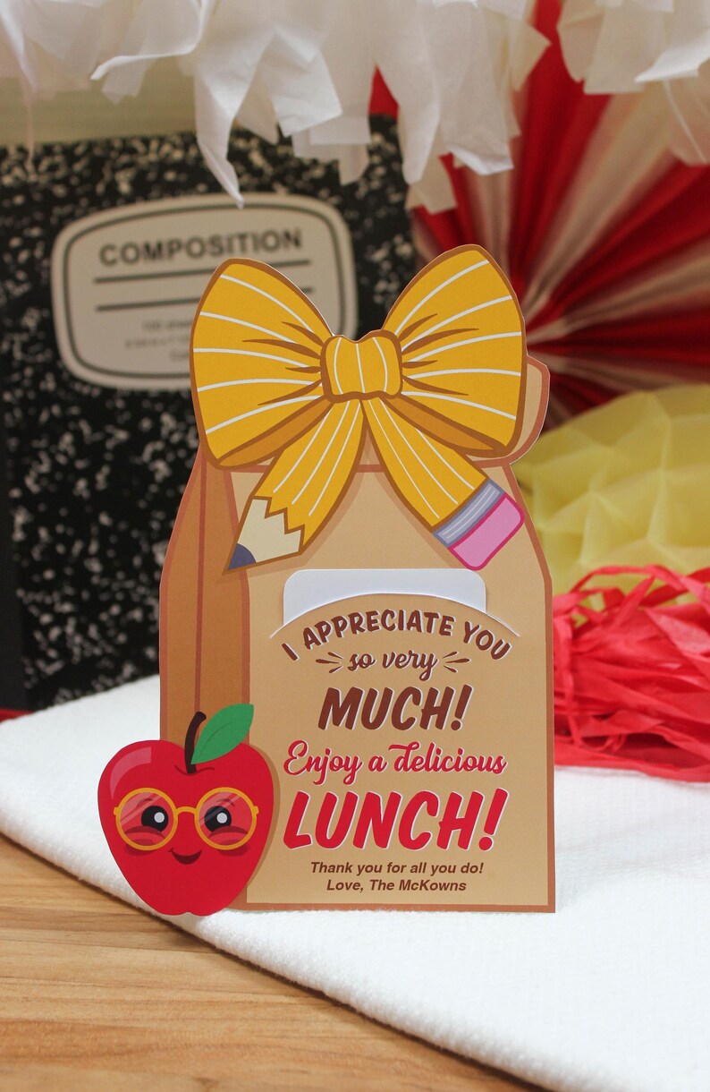 May include: A brown paper bag with a yellow bow and a red apple with glasses. The bag has a pencil and a ruler sticking out of it. The text on the bag reads "I appreciate you so very much! Enjoy a delicious lunch! Thank you for all you do! Love, The McKowns."