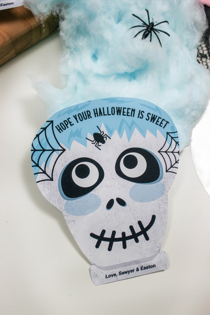 Puede incluir: Una tarjeta de Halloween azul y blanca con una cara de calavera de dibujos animados y el texto "Espero que tu Halloween sea dulce. Con cari&ntilde;o, Sawyer & Easton".