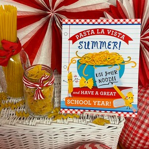 May include: A white wicker basket with red and white striped paper lanterns and a sign that says "Pasta La Vista Summer! Use your noodle and have a great school year!" The sign has a cartoon noodle and a cartoon bowtie pasta on it.