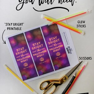 Op de afbeelding: Een bedrukbaar vel met "Stay Bright This Halloween Night"-etiketten met een paarse en roze achtergrond en een pompoenafbeelding. Het vel is omringd door glowsticks, een schaar, een stanleymes en een zak confetti.