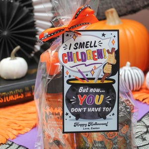 May include: A Halloween gift bag with a tag that reads "I smell children! But now... you don't have to!" The bag contains a pumpkin-scented hand sanitizer and a pumpkin carving candle. The tag is decorated with a witch's cauldron and a broom.