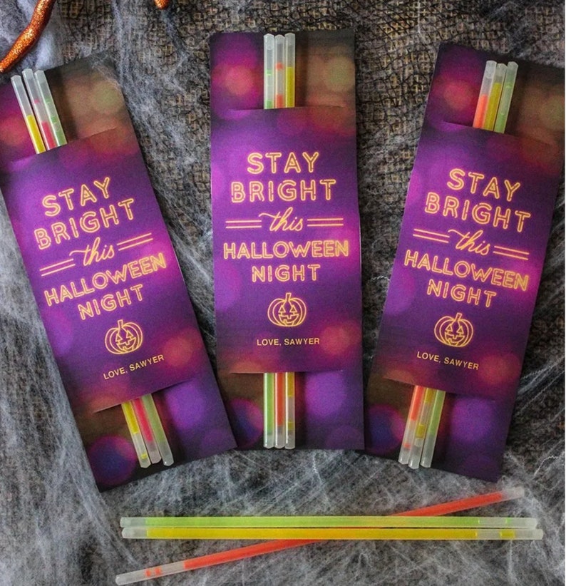 Op de afbeelding: Drie paarse en gouden Halloween-feesttasjes met glowsticks erin. Elk tasje heeft de tekst "Stay Bright =this= Halloween Night" en "Love, Sawyer".