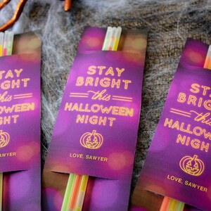 Op de afbeelding: Drie paarse kaarten met glowsticks erin. De kaarten hebben een Halloween-thema en zeggen "Stay Bright this Halloween Night" met een pompoenafbeelding en "Love, Sawyer".