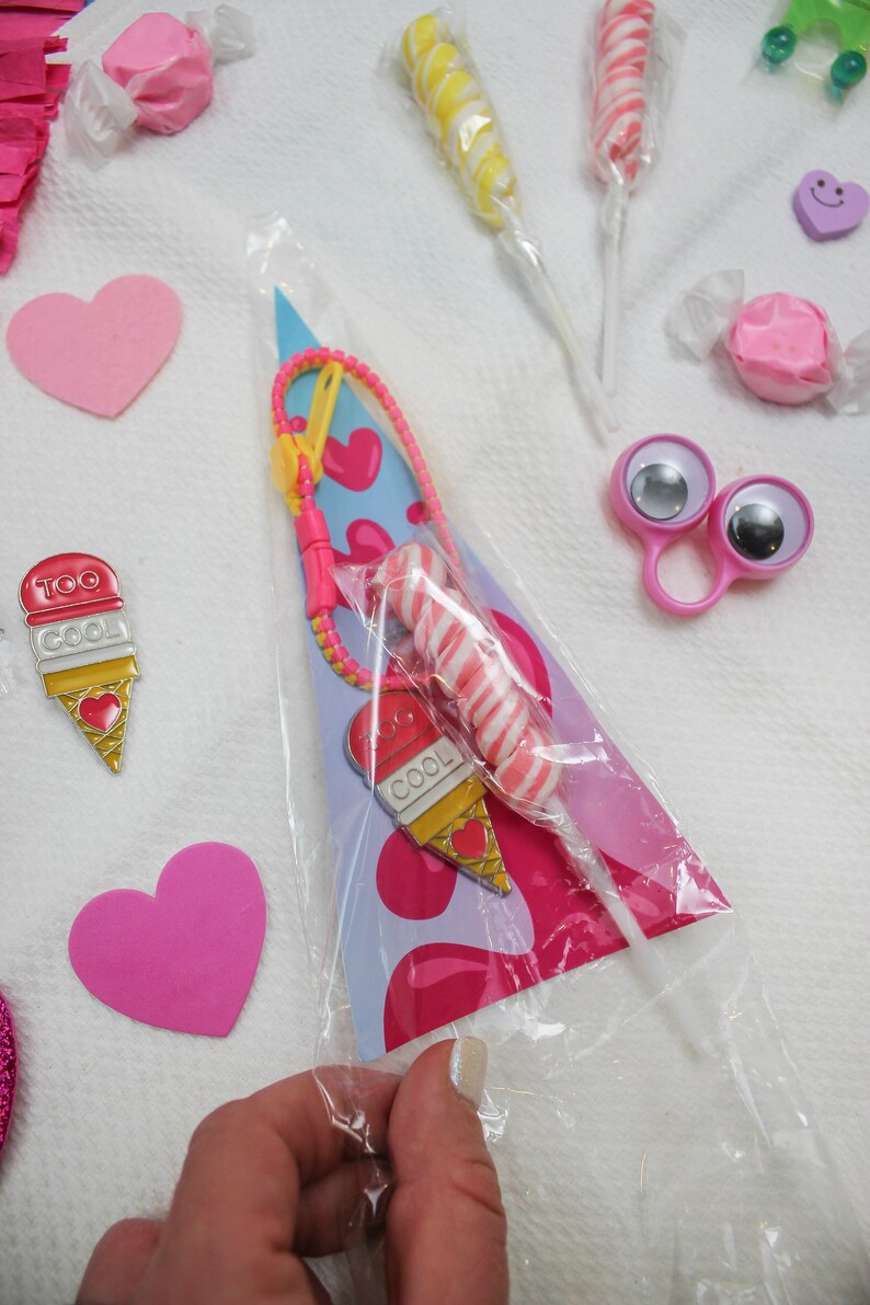Peut inclure: Un sac en plastique transparent rempli de cadeaux de f&ecirc;te. Le sac contient une sucette en spirale rose et blanche, un charme en forme de cornet de glace rose et blanc, un charme en forme de c&oelig;ur bleu et blanc et un bracelet en perles jaunes et roses. Le sac est pos&eacute; sur une surface blanche.