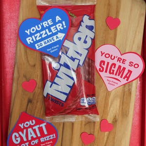 May include: A red wooden tray with a package of Twizzlers candy and several heart-shaped paper notes. The notes have different messages, including "You're a rizzler! So have a...", "You're so sigma", and "You gyatt a lot of rizz!"