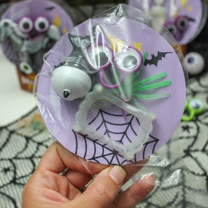 May include: A purple and white Halloween party favor bag with a spider, googly eyes, fangs, and a spider web design. The bag is filled with small Halloween toys.