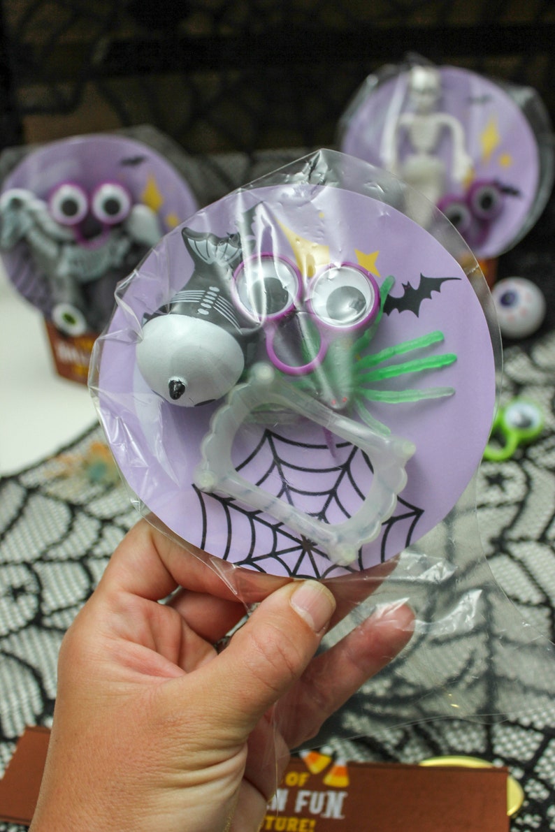 May include: A purple and white Halloween party favor bag with a spider, googly eyes, fangs, and a spider web design. The bag is filled with small Halloween toys.