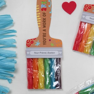 May include: A paintbrush-shaped gift tag with the text "YOU'RE A WORK OF ART!" and "Your Friend, Easton." The brush bristles are rainbow-colored candy sticks. Red heart-shaped decorations and blue paper streamers are also present.