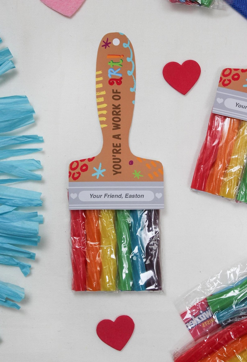 May include: A paintbrush-shaped gift tag with the text "YOU'RE A WORK OF ART!" and "Your Friend, Easton." The brush bristles are rainbow-colored candy sticks. Red heart-shaped decorations and blue paper streamers are also present.