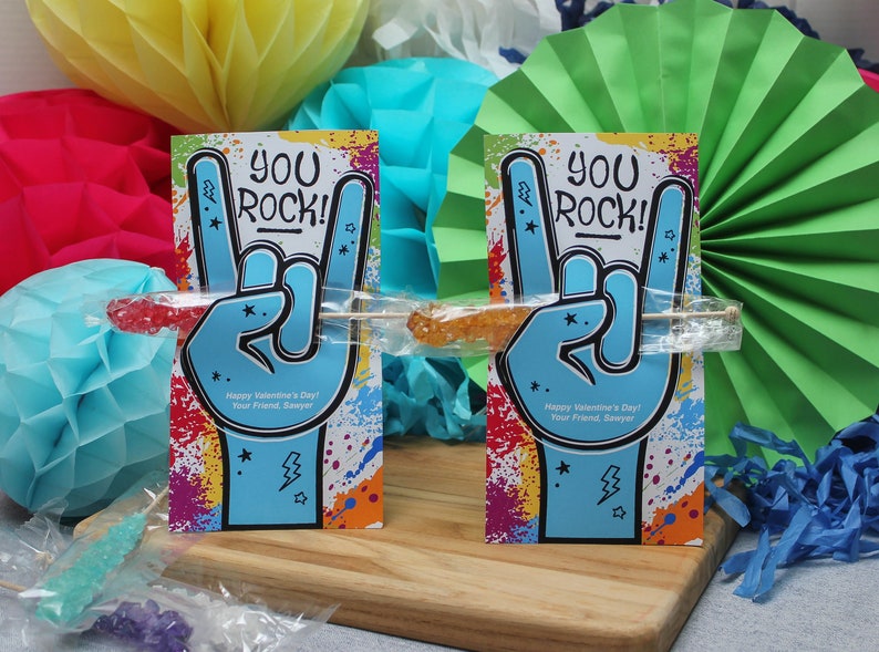 Pode incluir: Dois cart&otilde;es coloridos com o texto "You Rock!" e um gesto de m&atilde;o de rock and roll. Cada cart&atilde;o tem um bast&atilde;o de caramelo anexado.