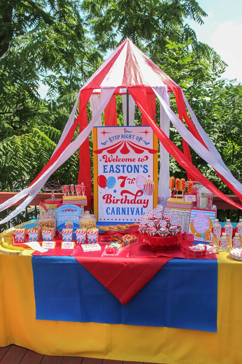 Peut inclure: D&eacute;coration de f&ecirc;te d'anniversaire sur le th&egrave;me du carnaval, avec des couleurs rouge, blanc et bleu. La table est orn&eacute;e de pop-corn, de petits g&acirc;teaux et d'une pancarte indiquant "Bienvenue au 7e anniversaire de carnaval d'Easton". Une structure de tente &agrave; rayures rouges et blanches est au-dessus.