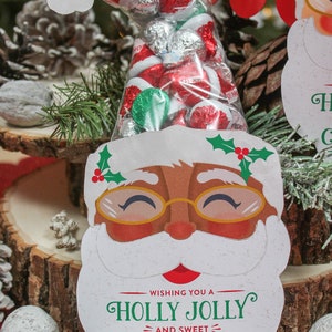 May include: A white Santa Claus face cutout with a red nose and black eyes. The cutout has a red and green holly sprig on top and the words "Wishing you a Holly Jolly and Sweet Christmas! Love, Sawyer and Easton" at the bottom. A clear plastic bag filled with red and silver foil-wrapped candies is tucked into the cutout.