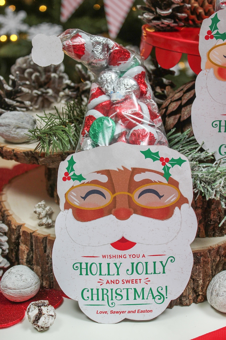 May include: A white Santa Claus face cutout with a red nose and black eyes. The cutout has a red and green holly sprig on top and the words "Wishing you a Holly Jolly and Sweet Christmas! Love, Sawyer and Easton" at the bottom. A clear plastic bag filled with red and silver foil-wrapped candies is tucked into the cutout.