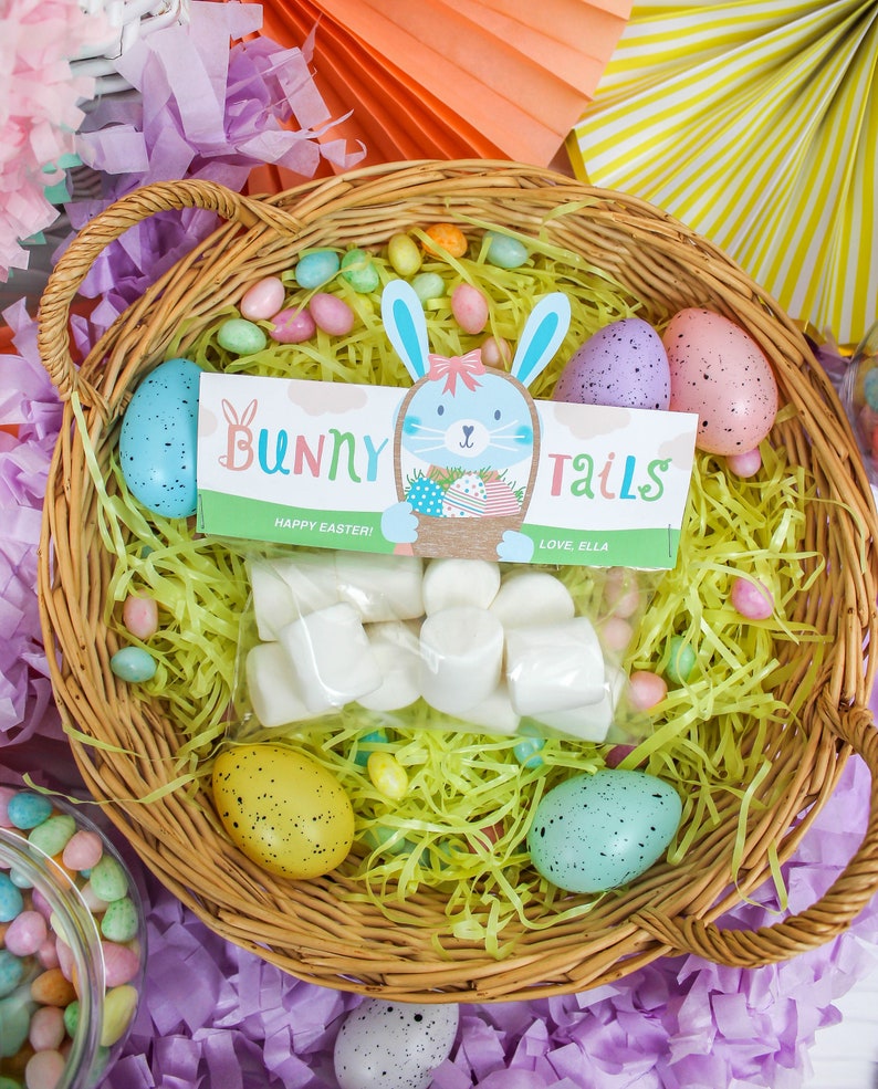 May include: A wicker basket filled with yellow shredded paper, pastel-colored jelly beans, and a bag of white marshmallows. There is a printable tag with the text "Bunny Tails" and a cute bunny illustration. The tag also says "Happy Easter!" and "Love, Ella".