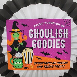 May include: A pink and orange Halloween sign with a witch holding a spoon, a mug with a monster face, and a bucket of popcorn. The sign reads "Proud Purveyor of Ghoulish Goodies Spooktacular Snacks and Tricky Treats".