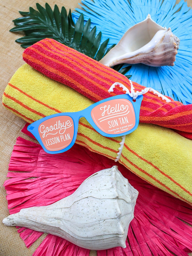 May include: A pair of blue sunglasses with the text "Goodbye Lesson Plan" on one lens and "Hello Sun Tan" on the other lens. The sunglasses are placed on top of a rolled up yellow and orange striped beach towel.