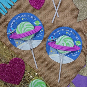Op de afbeelding: Twee groene en witte swirl-lollipops met een blauwe en roze ruimte-thema Valentijnskaart die aan elk is bevestigd. De kaarten hebben de tekst "Valentijn, je bent niet van deze wereld!" en "Ik ben zo blij dat we dezelfde ruimte delen!" met een roze ring rond een groene en witte swirl-lollipop.