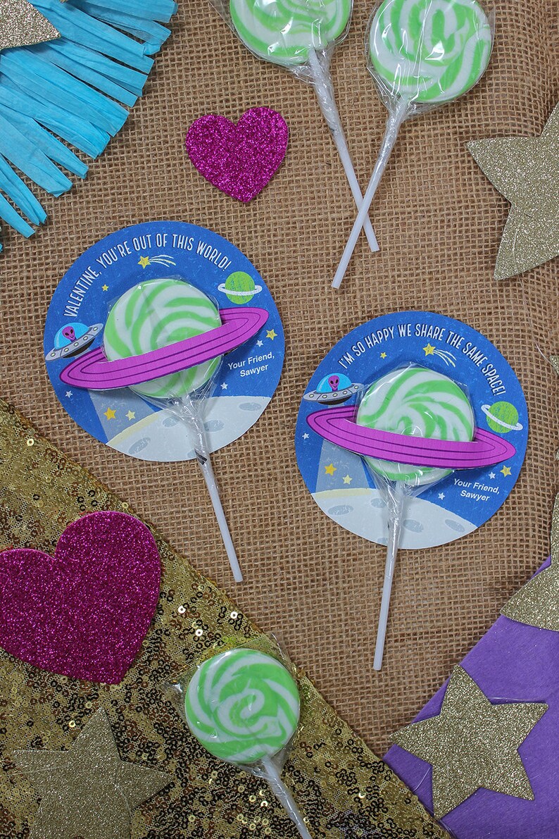 Op de afbeelding: Twee groene en witte swirl-lollipops met een blauwe en roze ruimte-thema Valentijnskaart die aan elk is bevestigd. De kaarten hebben de tekst "Valentijn, je bent niet van deze wereld!" en "Ik ben zo blij dat we dezelfde ruimte delen!" met een roze ring rond een groene en witte swirl-lollipop.