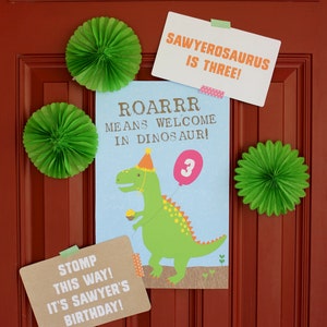 May include: A red door decorated with green paper fans and signs for a dinosaur themed birthday party. The signs say "Sawyerosaurus is Three!" and "Roarrr means welcome in dinosaur!" with a cartoon dinosaur holding a balloon that says "3". Another sign says "Stomp this way! It's Sawyer's birthday!"