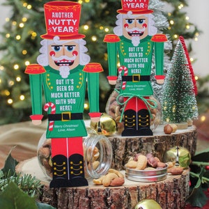 May include: Two nutcracker figurines holding jars of mixed nuts. The nutcrackers are dressed in red, green, and black with a white hat. The jars are decorated with a red and white candy cane and a red ribbon. The text on the nutcrackers reads "Another Nutty Year" and "But it's been so much better with you here!"