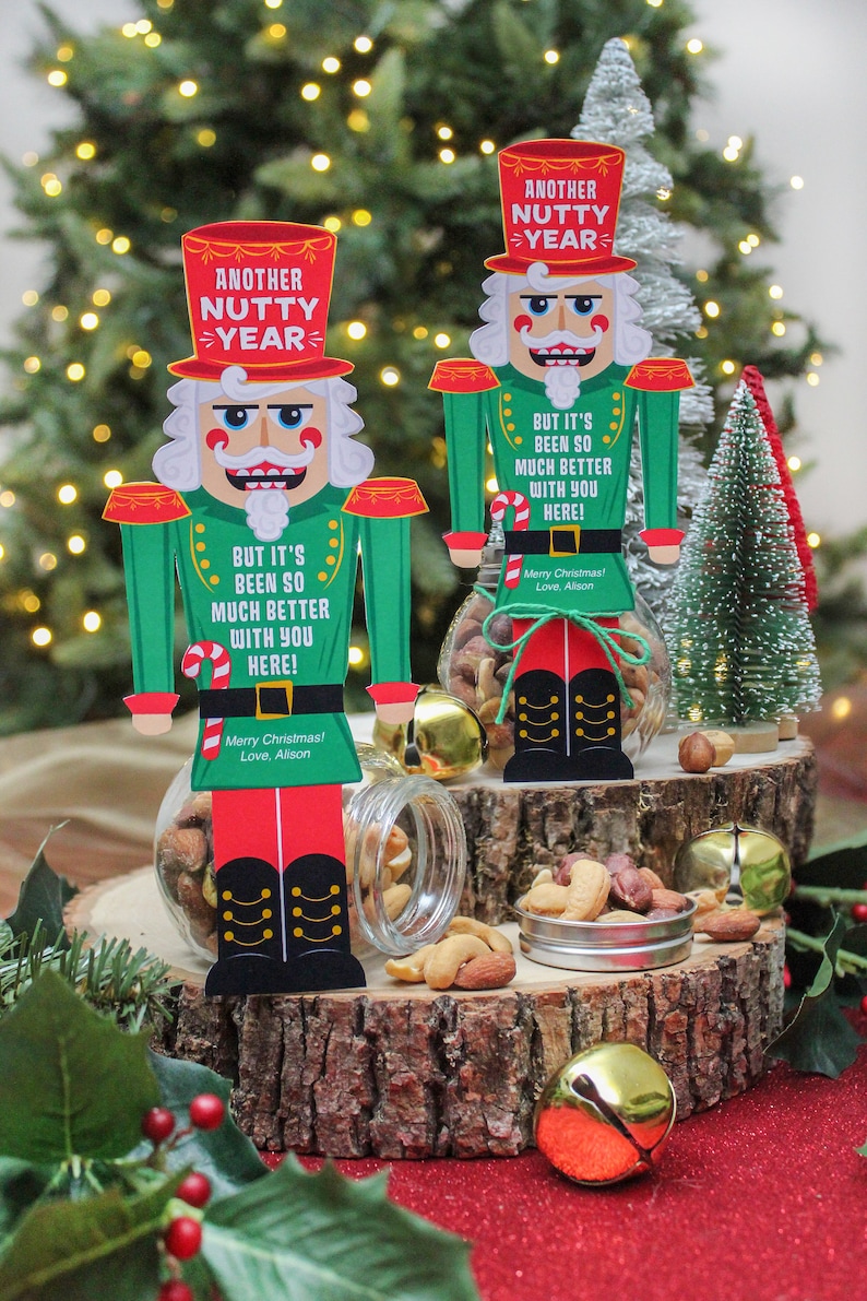 May include: Two nutcracker figurines holding jars of mixed nuts. The nutcrackers are dressed in red, green, and black with a white hat. The jars are decorated with a red and white candy cane and a red ribbon. The text on the nutcrackers reads "Another Nutty Year" and "But it's been so much better with you here!"