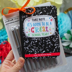 Könnte beinhalten: Ein Geschenkset für die Schuleinführung mit einem schwarzen Notizbuch mit dem Text "TAKE NOTE... IT'S GOING TO BE A GREAT SCHOOL YEAR!" und einer Packung silberner Stifte. Ein orangefarbenes Band bindet das Set zusammen.
