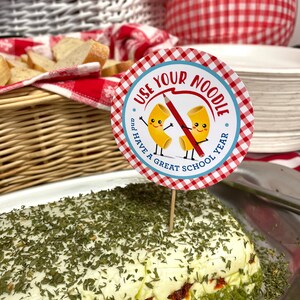 May include: A white and green layered dip with a red sauce in between the layers. The dip is topped with green herbs. A red and white checkered circle with the text "Use Your Noodle and Have a Great School Year" is on a toothpick in front of the dip.