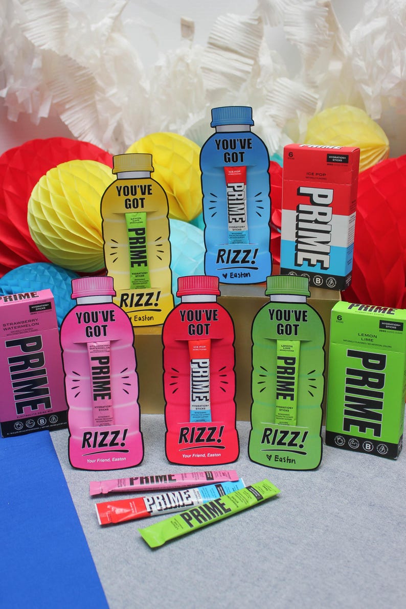 May include: Six colourful cardboard boxes shaped like water bottles with the text "You've Got Prime" and "Rizz!" on them. Each box contains a single-serving hydration stick. The boxes are arranged in a semi-circle on a grey surface. There are two boxes of Prime hydration sticks in the foreground.
