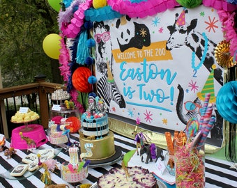 May include: A birthday party table decorated with a zebra, giraffe, and panda theme. The table has a black and white striped tablecloth and is decorated with colorful balloons, streamers, and paper decorations. There are several cakes, cookies, and other treats on the table. The banner says "Welcome to the Zoo Easton is Two!"