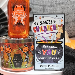 May include: A Halloween-themed candle with a pumpkin carving design and skeletons dancing. The candle is in a clear glass jar with a black lid. A card with the text "I smell children! But now... you don't have to! Happy Halloween! Love, Easton" is in the foreground.