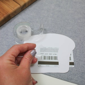 May include: A white gift card with a barcode and the text "CARD# 6207 9897 6788". The card is being taped to a white paper shape.