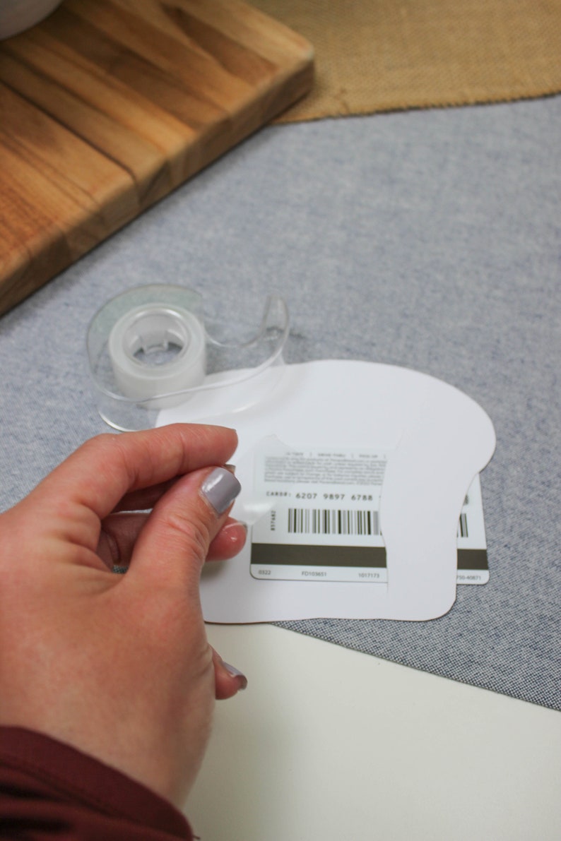 May include: A white gift card with a barcode and the text "CARD# 6207 9897 6788". The card is being taped to a white paper shape.