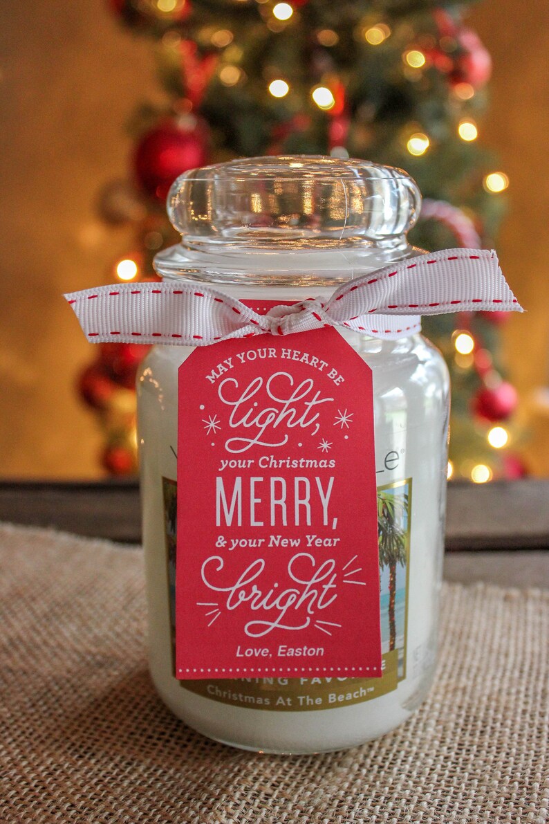 May include: A white candle jar with a red and white ribbon and a red tag that says "May your heart be light, your Christmas Merry, & your New Year bright. Love, Easton. Christmas At The Beach."