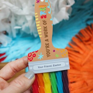 May include: A brown paintbrush-shaped card with the text "YOU'RE A WORK OF ART!" and "Your Friend, Easton." The brush bristles are rainbow-colored candy. The background has white, blue, and orange decorations.