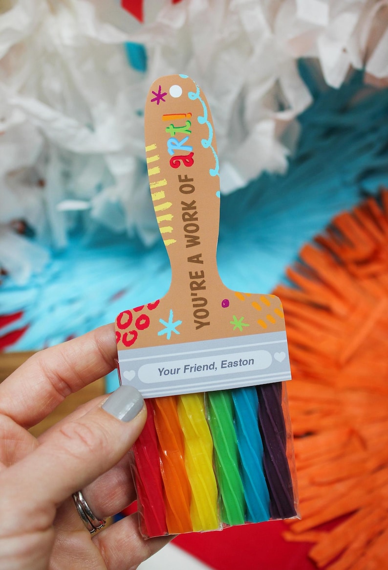 May include: A brown paintbrush-shaped card with the text "YOU'RE A WORK OF ART!" and "Your Friend, Easton." The brush bristles are rainbow-colored candy. The background has white, blue, and orange decorations.